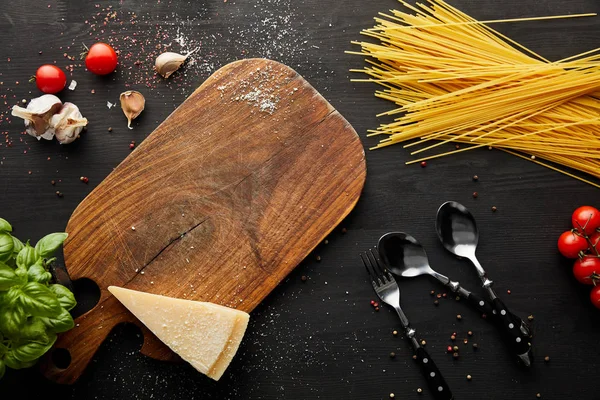 Vista superior de los ingredientes de pasta boloñesa, tabla de cortar y cubiertos sobre fondo de madera negro - foto de stock