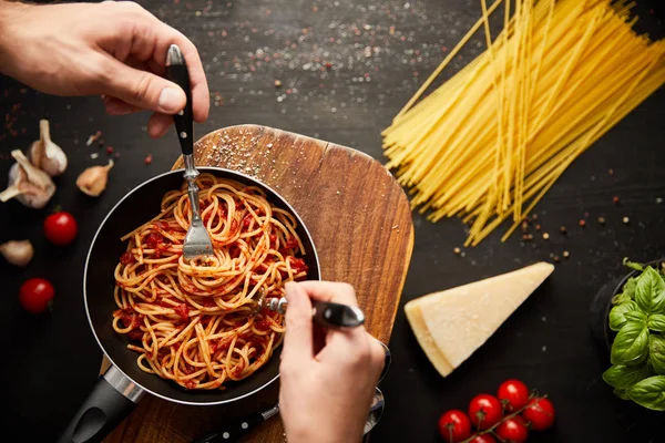 Cropped view of couple eating tasty bolognese pasta from frying pan near ingredients on black wooden background — Stock Photo