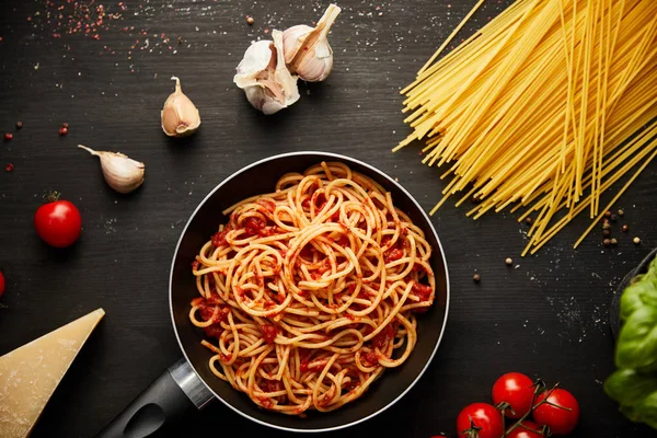 Top view of tasty bolognese pasta in frying pan near ingredients on black wooden background — Stock Photo