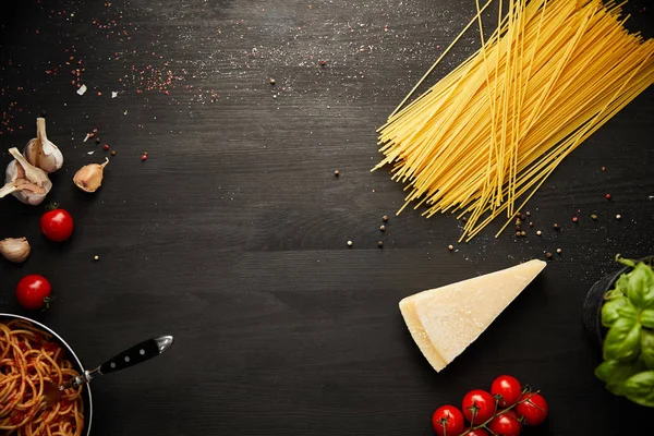 Top view of tasty bolognese pasta in frying pan near ingredients on black wooden background with copy space — Stock Photo