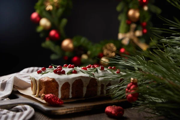 Selective focus of traditional Christmas cake with cranberry near Christmas wreath with baubles on wooden table isolated on black — Stock Photo