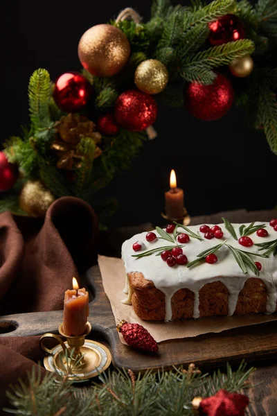 Gâteau de Noël traditionnel avec canneberge près de couronne de Noël avec des boules et des bougies sur table en bois isolé sur noir — Photo de stock