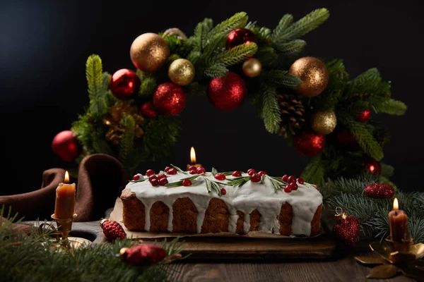 Selective focus of traditional Christmas cake with cranberry near Christmas wreath with baubles on wooden table isolated on black — Stock Photo