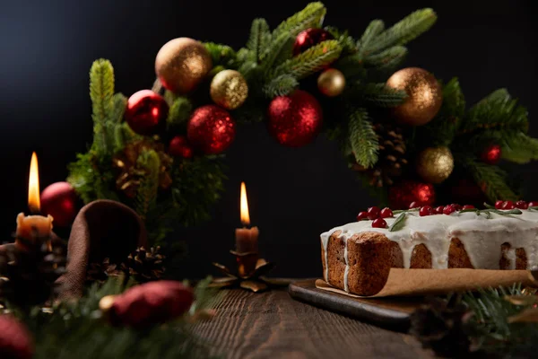 Traditional Christmas cake with cranberry near Christmas wreath with baubles and burning candles on wooden table isolated on black — Stock Photo