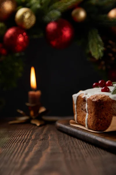 Selective focus of traditional Christmas cake with cranberry near Christmas wreath with baubles and burning candle on wooden table isolated on black — Stock Photo