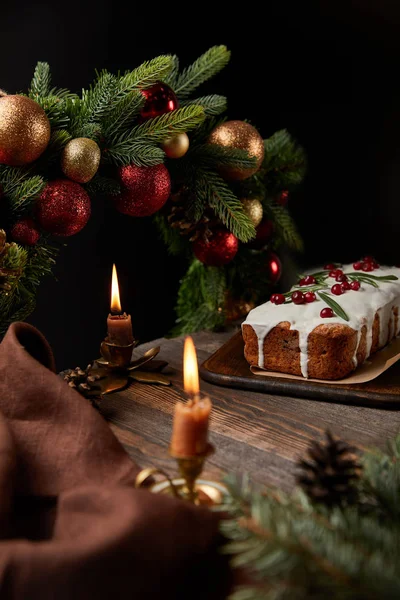 Selective focus of traditional Christmas cake with cranberry near Christmas wreath with baubles and burning candles on wooden table isolated on black — Stock Photo