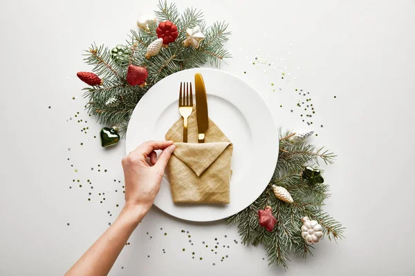 Vue recadrée d'une femme tenant une serviette sur une assiette blanche avec des couverts près d'une branche festive d'arbre de Noël et des confettis sur fond blanc — Photo de stock