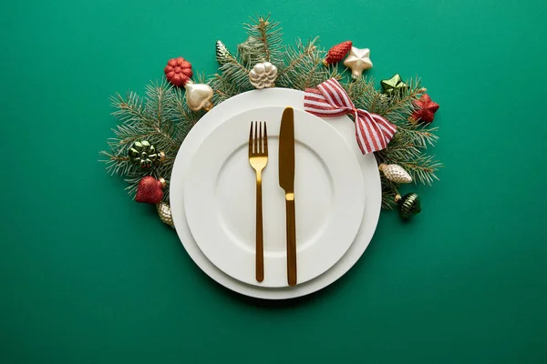 Top view of white plates with cutlery near festive Christmas tree branch with baubles on green background — Stock Photo