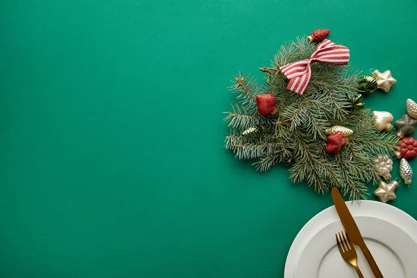 Vista dall'alto di piatto bianco con posate vicino a ramo di albero di Natale festivo con bagattelle su sfondo verde con spazio copia — Foto stock