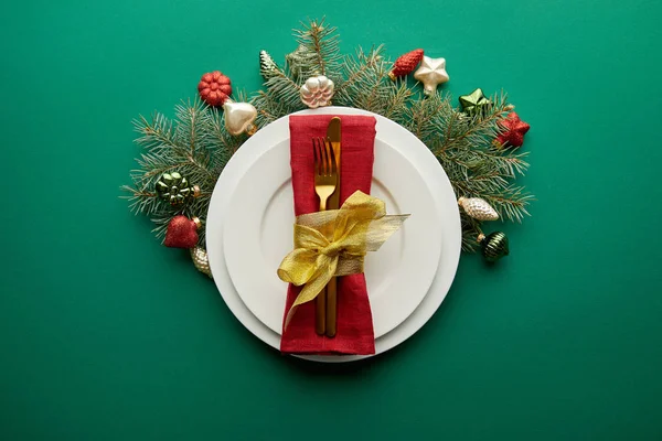 Vista dall'alto di piatto bianco con tovagliolo, posate vicino a ramo di albero di Natale festivo con bagattelle su sfondo verde — Foto stock