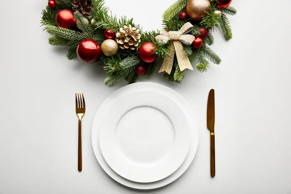 Top view of white plates with golden cutlery near festive Christmas wreath with baubles on white background — Stock Photo