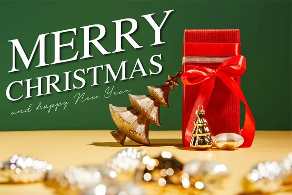 Selective focus of red gift box and decorative Christmas tree with golden baubles on green background with Merry Christmas and happy new year illustration — Stock Photo