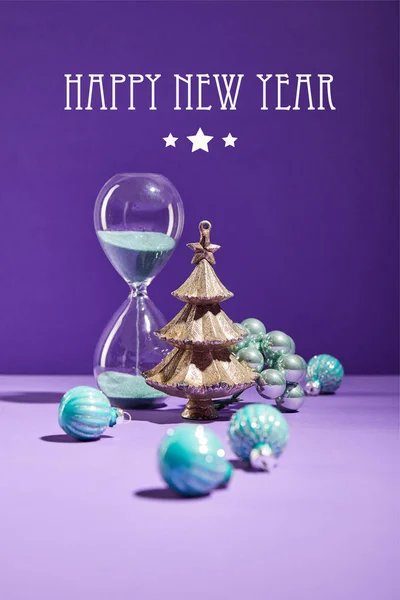 Selektiver Schwerpunkt der dekorativen Weihnachten in der Nähe von blauen Christbaumkugeln und Sanduhr auf lila Hintergrund mit frohem neuen Jahr Schriftzug — Stockfoto