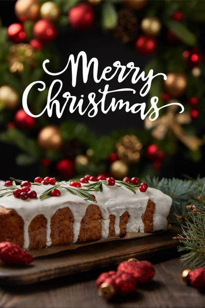 Selective focus of traditional Christmas cake with cranberry near Christmas wreath with baubles on wooden table with Merry Christmas illustration — Stock Photo