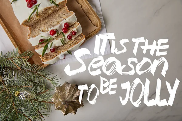 Верхний вид на традиционный рождественский торт с клюквой рядом безделушки и сосны на белом столе с ним сезон, чтобы быть веселым буквы — стоковое фото