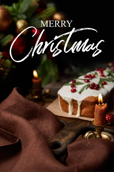 Selective focus of traditional Christmas cake with cranberry near burning candles on wooden table with Merry Christmas illustration — Stock Photo
