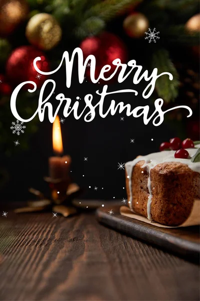 Selective focus of traditional Christmas cake with cranberry near Christmas wreath with baubles and burning candle on wooden table with Merry Christmas illustration — Stock Photo