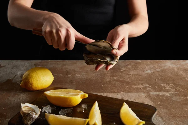 Vue recadrée de la femme tenant un couteau lors de l'ouverture huître près de citrons isolés sur noir — Photo de stock