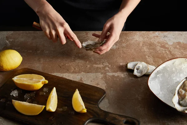 Vue recadrée du chef tenant un couteau lors de l'ouverture de l'huître près de citrons isolés sur noir — Photo de stock