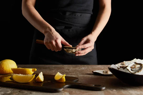 Vista recortada del cuchillo de chef mientras se abre la ostra cerca de limones en la tabla de cortar aislado en negro - foto de stock