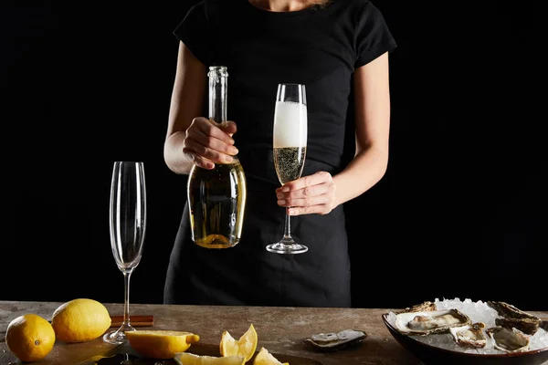 Vista cortada de mulher segurando vidro e garrafa com espumante vinho neae ostras em tigela com gelo isolado em preto — Fotografia de Stock