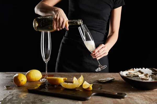 Vista recortada de la mujer vertiendo vino espumoso en copa de champán cerca de limones y ostras aislados en negro - foto de stock