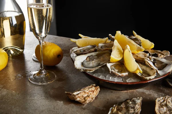 Messa a fuoco selettiva di ostriche e limoni in ciotola con ghiaccio vicino allo spumante in bicchiere di champagne isolato su nero — Foto stock