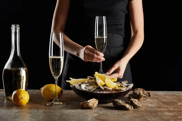 Vista cortada de mulher segurando vidro com vinho espumante perto de ostras e limões em tigela com gelo isolado em preto — Fotografia de Stock