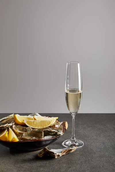 Вкусные устрицы и лимоны в миске со льдом возле бокала шампанского с игристым вином, изолированным на сером — стоковое фото