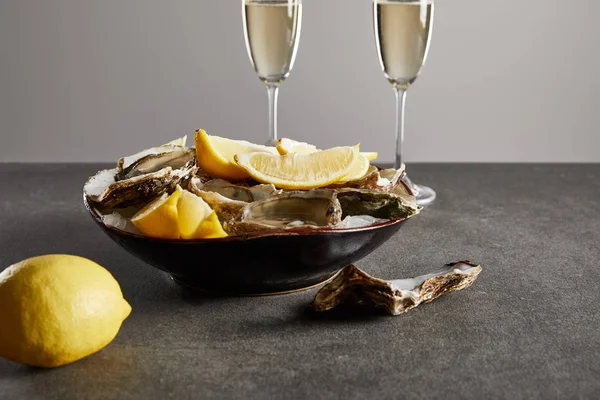 Messa a fuoco selettiva di deliziose ostriche e limoni in ciotola vicino a bicchieri di champagne con spumante isolato su grigio — Foto stock