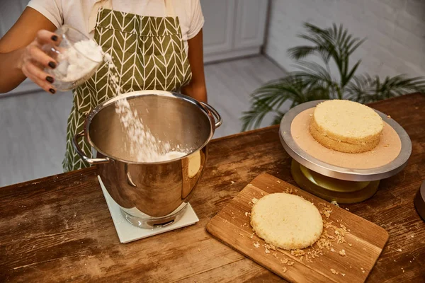 Vista cortada de confeiteiro adicionando farinha na tigela ao lado de camadas de bolo na mesa — Fotografia de Stock