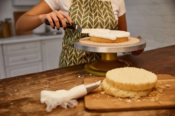 Cropped view of confectioner with spatula putting cream on sponge cake layer — Stock Photo