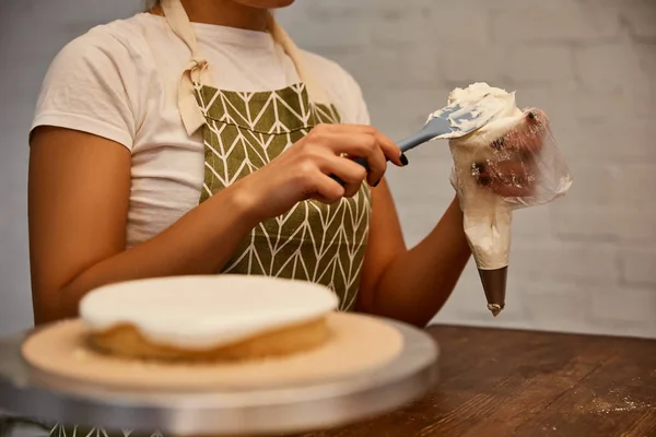 Vista cortada de confeiteiro colocando creme no saco de pastelaria ao lado da camada de bolo na mesa — Fotografia de Stock