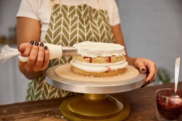 Vista ritagliata del pasticciere che mette la crema dalla borsa della pasticceria sul pan di Spagna — Foto stock