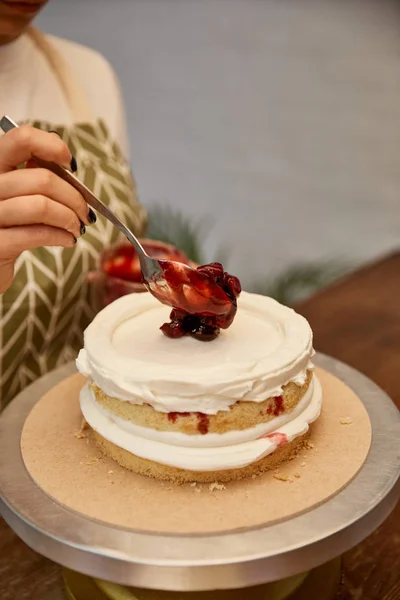 Vista recortada del confitero con cuchara poniendo mermelada de bayas en la torta con crema - foto de stock