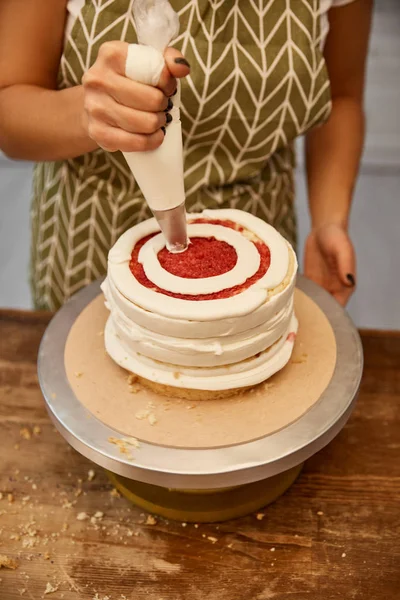 Vista ritagliata del pasticciere aggiungendo panna dal sacchetto di pasticceria sulla torta — Foto stock