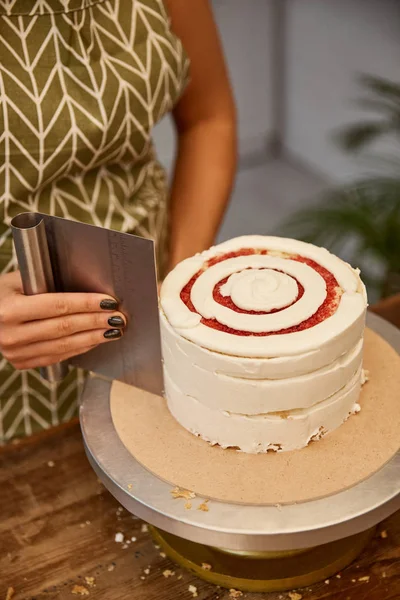 Cropped view of confectioner aligning cream with scraper on sponge cake — Stock Photo