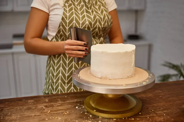 Vista recortada del confitero con raspador alineando crema dulce en la torta - foto de stock