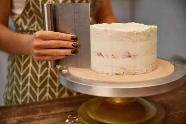 Cropped view of confectioner using scraper on tasty cream on sponge cake — Stock Photo