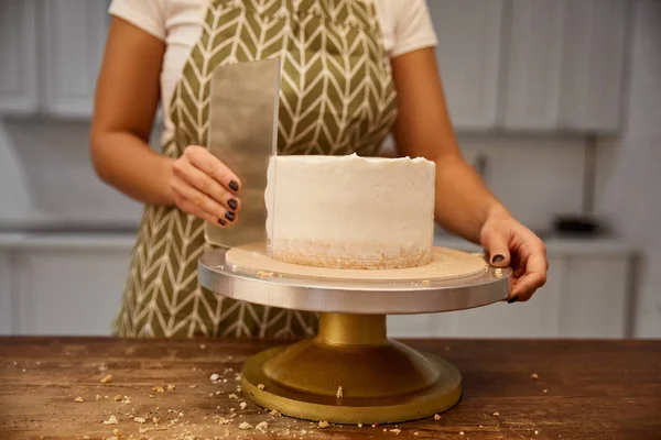 Cropped view of female confectioner aligning cream with scraper on sponge cake — Stock Photo