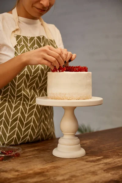 Vista ritagliata del pasticcere sorridente che decora la torta con ribes rosso dolce — Foto stock