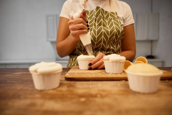 Abgeschnittene Ansicht des Konditors mit Teigtasche, die Sahne auf Cupcakes gießt — Stockfoto