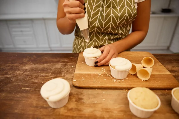 Cropped view of confectioner pouring cream on cupcakes beside waffle cones on cutting board — Stock Photo