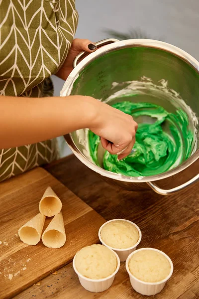 Vista cortada de confeiteiro misturando creme doce ao lado de cupcakes e cones de waffle na mesa — Fotografia de Stock