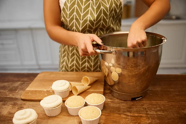 Vista ritagliata del pasticciere che fa la crema in ciotola accanto a cupcake freschi e tavolo coni di cialde — Foto stock