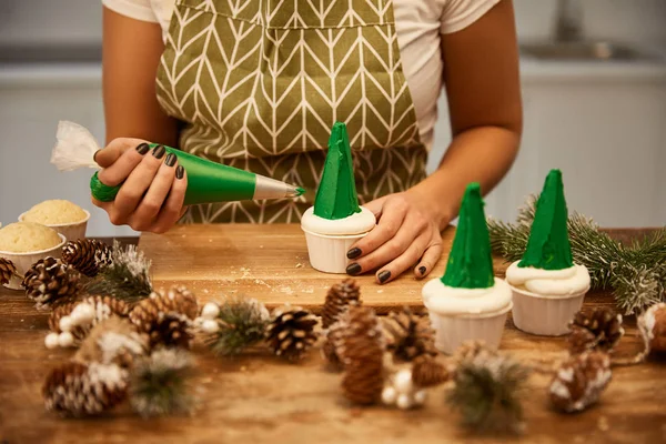 Vista cortada de confeiteiro decorando cupcake com saco de pastelaria ao lado de cones de pinho na mesa — Fotografia de Stock