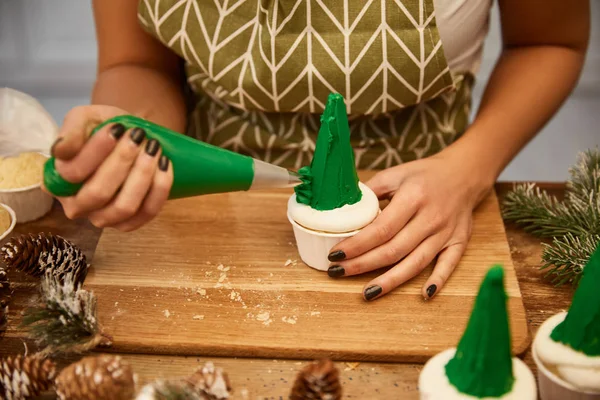Cropped view of confectioner decorating cupcakes with cream beside pine cones on table — Stock Photo