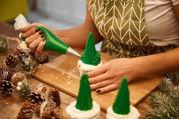 Vista ritagliata del pasticciere aggiungendo panna verde sui cupcake dell'albero di Natale accanto ai coni di abete rosso sul tavolo — Foto stock