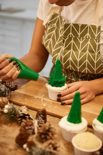 Cropped view of confectioner decorating Christmas tree cupcakes with sweet cream with spruce cones on wooden table — Stock Photo