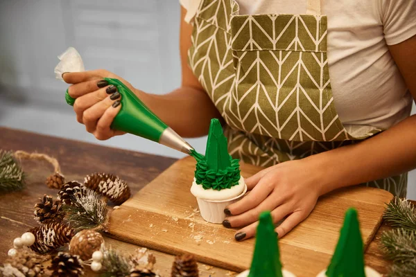 Vista cortada de confeiteiro adicionando creme saboroso em cupcakes de árvore de Natal com cones de abeto na mesa de madeira — Fotografia de Stock